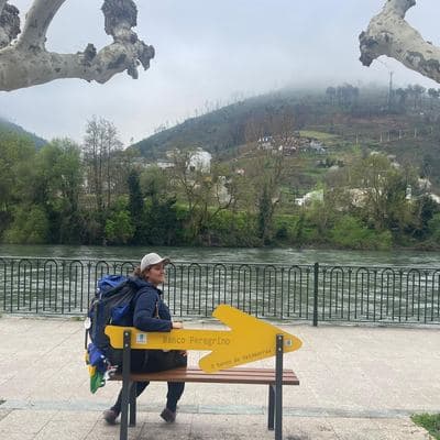 Vivi com mochila no banco peregrino, à beira do rio
